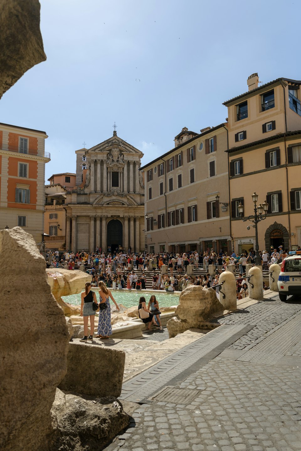 Des touristes se rassemblent autour d'une grande fontaine ornée sur une place de ville ensoleillée.